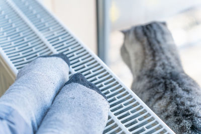 A woman warms her feet on the heater, a cat sits next to her and looks out the window.
