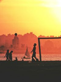 Silhouette people at beach by buildings against orange sky