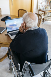 Back view of senior man in wheelchair using laptop at home