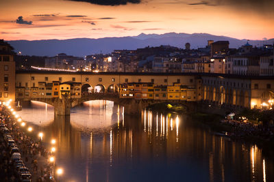 Illuminated bridge over river by buildings against sky during sunset