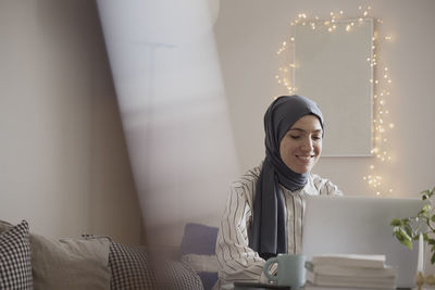 Smiling female freelancer wearing hijab while working on laptop in living room at home