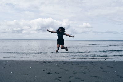 Woman with arms outstretched jumping at beach against cloudy sky