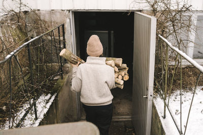 Rear view of woman carrying firewood while walking towards shed