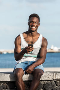 Portrait of smiling young man using mobile phone against sea
