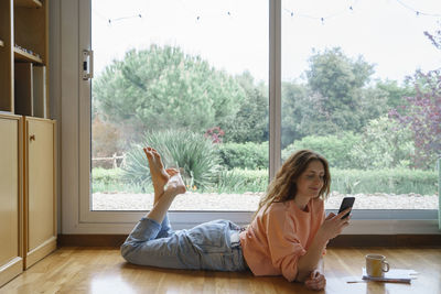 Young woman using phone while sitting on window at home