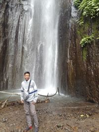 Full length of young man standing against waterfall in forest