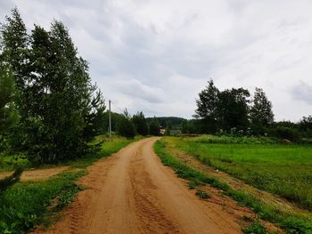 Dirt road along trees on field against sky