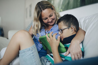 Smiling mother looking at disabled son using digital tablet while sitting on sofa