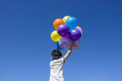 Low angle rear view of boy holding multi colored balloons against clear blue sky