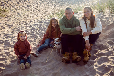 Family with a son and daughter and a dog sit on the sand in autumn