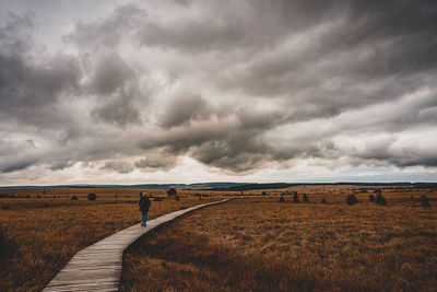 Woman walking on boardwalk amidst field against storm clouds