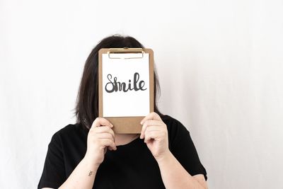 Woman hiding face with smile message on clipboard against wall