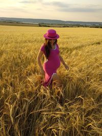 Pregnant woman wearing hat walking amidst crops on field during sunset