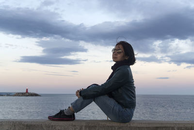 Side view of young woman sitting on beach against sky