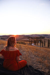 Rear view of woman sitting on field against sky during sunset