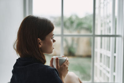 Side view of woman holding coffee cup against window