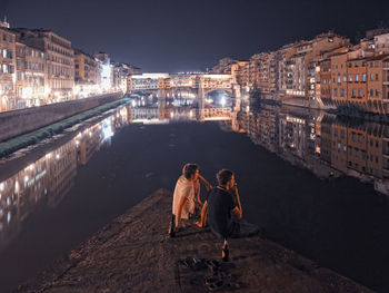 People sitting on bridge over river against sky in city at night