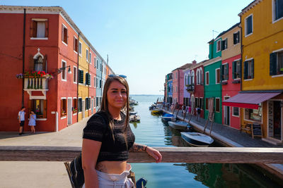 Portrait of young woman standing by canal against buildings in city