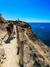 Rear view of people walking on rock by sea against clear blue sky