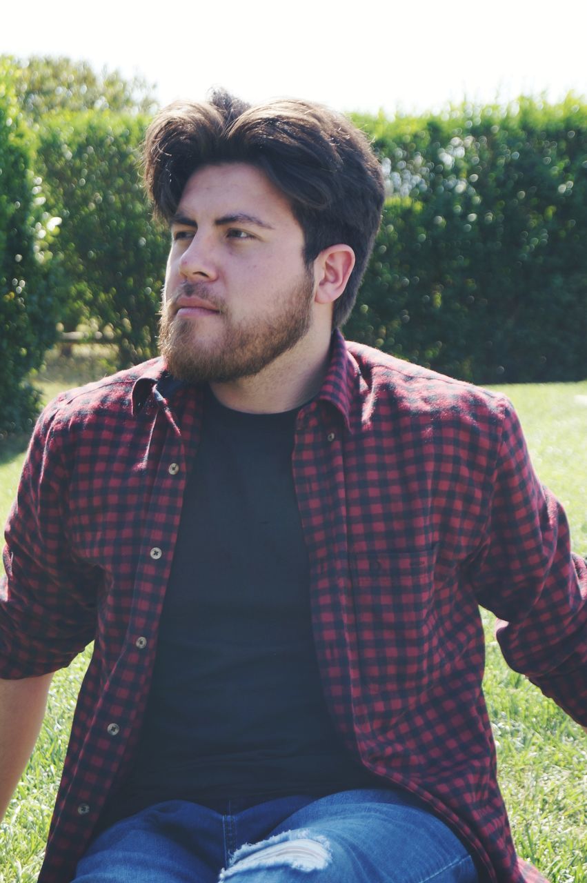 young adult, real people, young men, front view, one person, leisure activity, casual clothing, sitting, three quarter length, lifestyles, day, plant, nature, looking away, focus on foreground, looking, portrait, beard, outdoors, contemplation