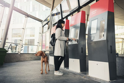 Side view of woman with dog using ticket machine at railroad station