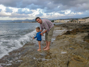 Portrait of father with son standing at beach against cloudy sky during sunset
