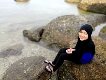 Portrait of young woman sitting on rock by sea