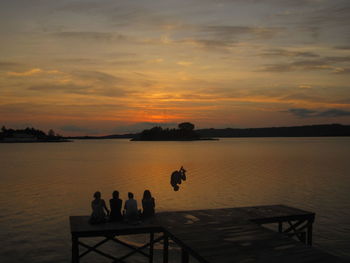 Friends looking at man jump while sitting on pier over lake during sunset