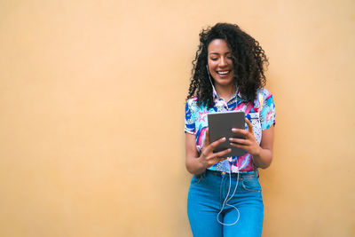 Smiling young woman using phone while standing against wall