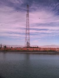 Electricity pylon by river against sky during sunset