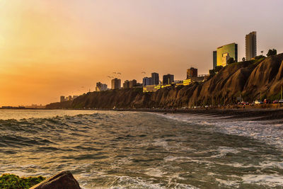 Sea by buildings against sky during sunset