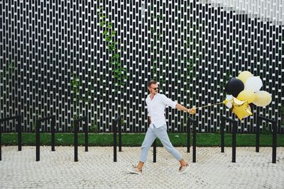 Fashionable man with balloons while walking on footpath against patterned wall