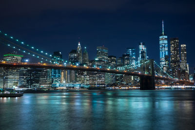 Illuminated brooklyn bridge over river against buildings at night
