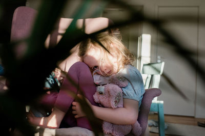 Young girl curled up on a chair looking tired holding her toy at home