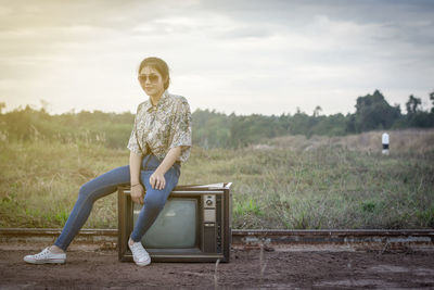 Portrait of young woman in sunglasses sitting on television against sky