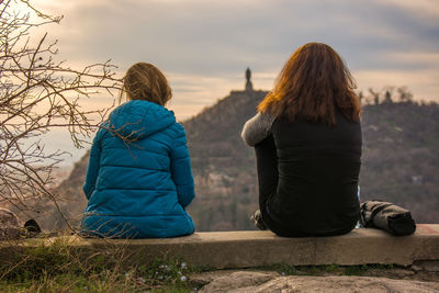 Rear view of female friends sitting on retaining wall against mountain