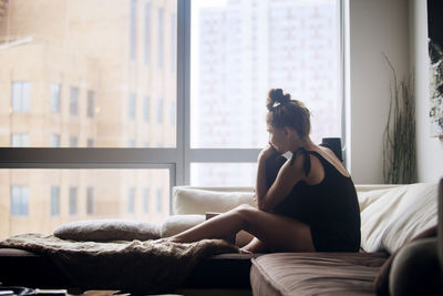 Woman looking through window while sitting on sofa at home