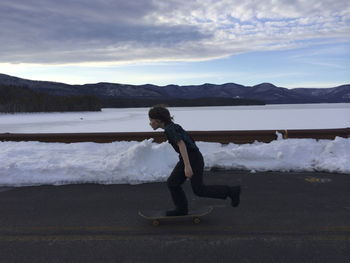 Full length side view of boy skateboarding on road by river during winter