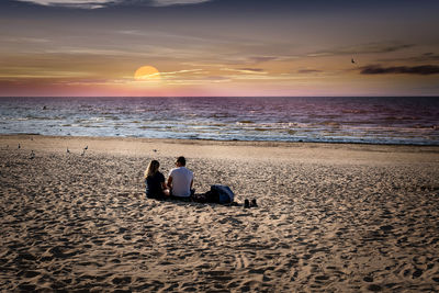 Friends sitting on shore at beach against sky during sunset