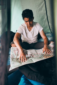 Young man looking at a map of mountain trails planning next trip on summer vacation sitting in tent