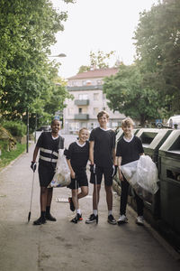Full length portrait of smiling boys standing with garbage bags by recycling bins