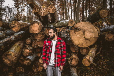 Bearded man standing against stacked logs in forest 