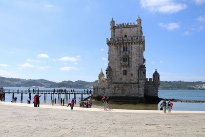 People on riverbank by belem tower against sky