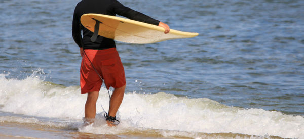 A man is standing at the waters edge walking into the ocean to go surfing.