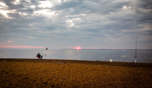 Man sitting at beach against sky during sunset