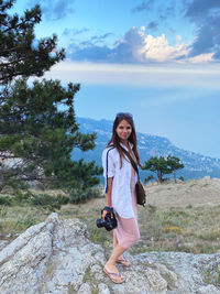 Portrait of young woman with camera standing against sky