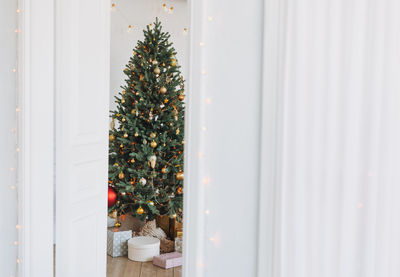 Decorated christmas tree in golden and white toys with the gift boxes from door at bright interior