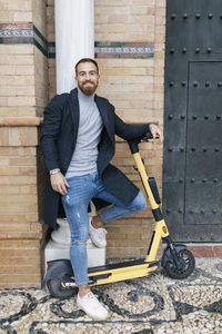Happy bearded man with push scooter leaning on column