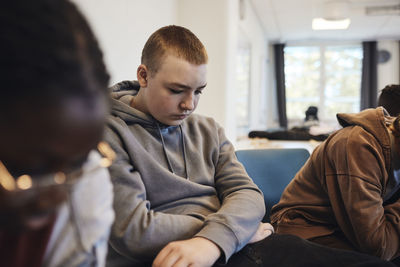 Depressed teenage boy with short hair sitting amidst female friends in group therapy at school