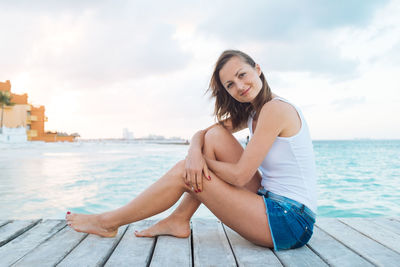 Portrait of smiling young woman sitting by sea against sky